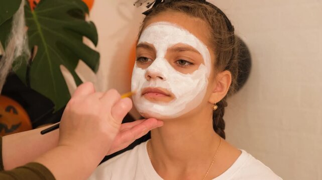 A cute teenage girl is getting ready for Halloween and doing face painting, with a skeleton painted on her face. October is a time of horror.