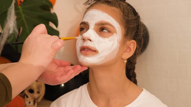 A cute teenage girl is getting ready for Halloween and doing face painting, with a skeleton painted on her face. October is a time of horror.