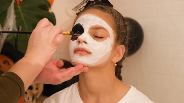 A cute teenage girl is getting ready for Halloween and doing face painting, with a skeleton painted on her face. October is a time of horror.