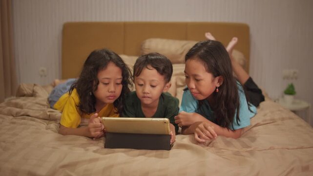 Three siblings, two sisters and a younger brother, lie on their stomachs on a bed while watching a movie on a tablet, capturing happy family bonding, childhood, and quality time at home.