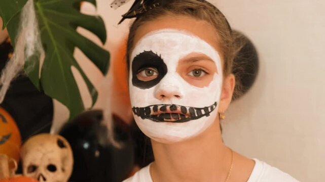 A cute teenage girl is getting ready for Halloween and doing face painting, with a skeleton painted on her face. October is a time of horror.
