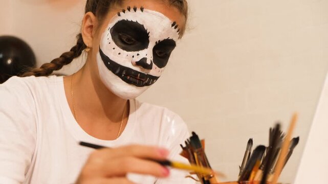 A cute teenage girl is getting ready for Halloween and doing face painting, with a skeleton painted on her face. October is a time of horror.