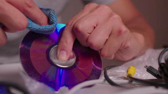 Hands polishing reflective compact disc on cluttered workbench with focused fingertip pressure, blue cloth movement, glossy highlights, and careful manual cleaning