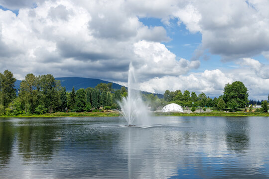 A tall fountain jet rises and reflects on the calm water of Lafarge Lake in Coquitlam, with a domed pavilion, green forest and mountains beneath a dramatic cloudy summer sky.