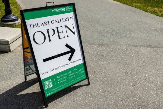 A green and white sandwich board reads The Art Gallery Is Open with a directional arrow and hours, outside the Evergreen Cultural Centre.