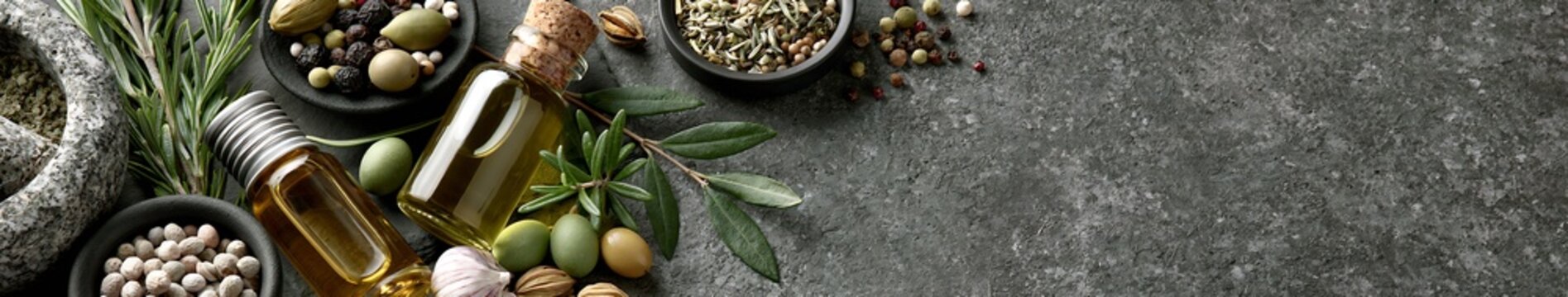Natural Skincare Olive oil bottles with fresh olives, rosemary, garlic and assorted spices on dark textured stone surface with copy space