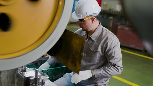 Factory worker wearing hard hat and goggles operates heavy machinery, inspecting a metal part with protective gloves in an industrial workshop beside a large press.