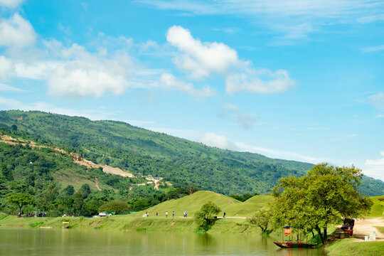 Scenic landscape featuring a calm river flowing past lush green hills and a mountain range under a bright blue sky with fluffy white clouds