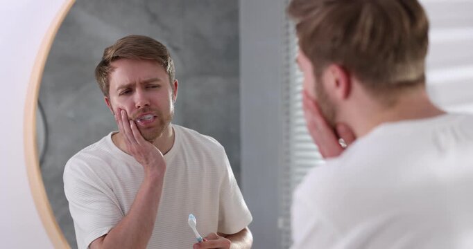 Man suffering from pain while brushing teeth near mirror in bathroom. Oral hygiene
