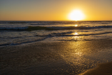 Obraz premium Golden sunlight reflects across gentle waves, illuminating wet sand in a warm, serene coastal sunset scene