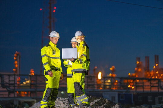 Three petrochemical engineers in yellow hi vis suits and white helmets reviewing turnaround plans on the laptop high above the site standing on a pale rock pile above an illuminated refinery