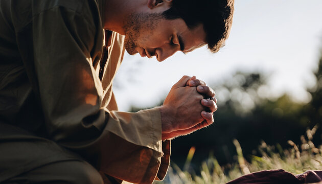 A man kneels outdoors with his hands clasped together, appearing to pray or meditate, with a natural landscape and trees in the background during sunset or sunrise.