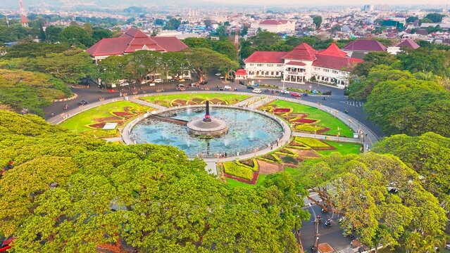 Sunrise illuminates MalangGolden hour bathes historic building and central monument. Circular pond reflects sky and structure. Lush greenery surrounds the scene. Distant trees frame the peaceful vista
