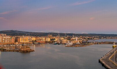 Fototapeta premium Vista panorámica de Civitavecchia ciudad costera y su puerto al atardecer, destacando la fortaleza histórica, barcos amarrados y un hermoso cielo pastel reflejado en aguas tranquilas.