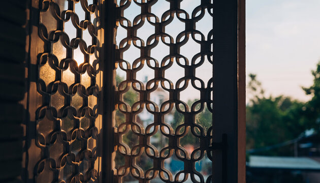 The image shows a decorative metal lattice window with an intricate geometric pattern, allowing sunlight to filter through and casting shadows inside a room during sunset.