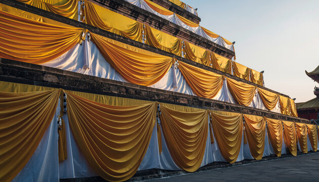 The image shows a multi-tiered structure decorated with yellow and white drapes, likely part of a festive or ceremonial event, set against a clear sky.