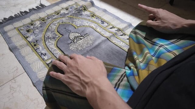 A Muslim man sitting on a prayer mat wearing a sarong and raising his hands for dua after performing salah