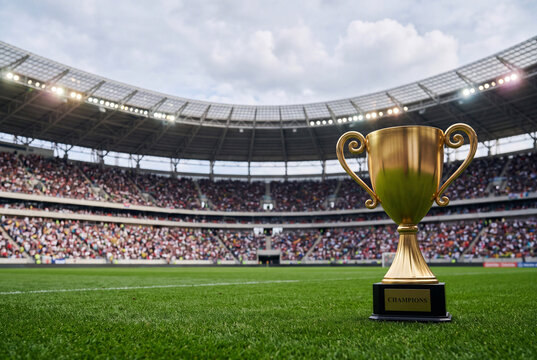 サッカースタジアムの芝生フィールドに置かれた金色トロフィーと観客席の熱気を捉えた勝利の象徴写真