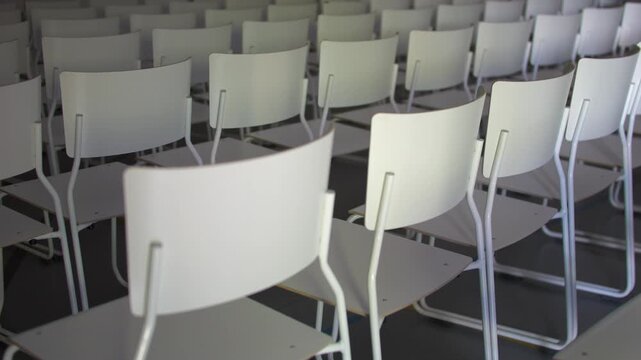 Rows of empty white chairs in modern auditorium conference room with smooth camera zoom.
