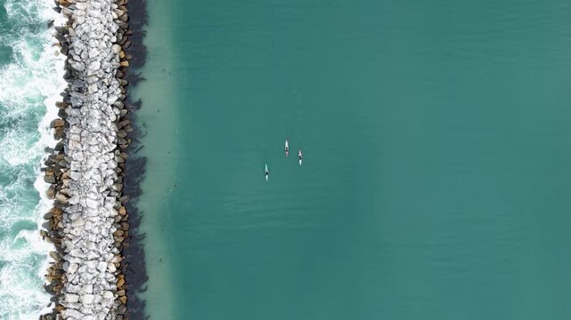 High altitude drone shot straight down on three kayakers gliding alongside the jetty at Dana Point Harbor. The contrast of the Pacific Ocean on one side and the calm harbor on the other is stunning
