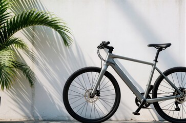 Fototapeta premium Grey hybrid bicycle standing against clean white wall with green palm leaves shadow ideal for urban mobility marketing and banner