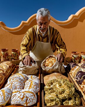 Mature Hispanic man placing fresh conchas and pan dulce into paper bags at a traditional bakery for Hispanic Heritage Month and food storytelling
