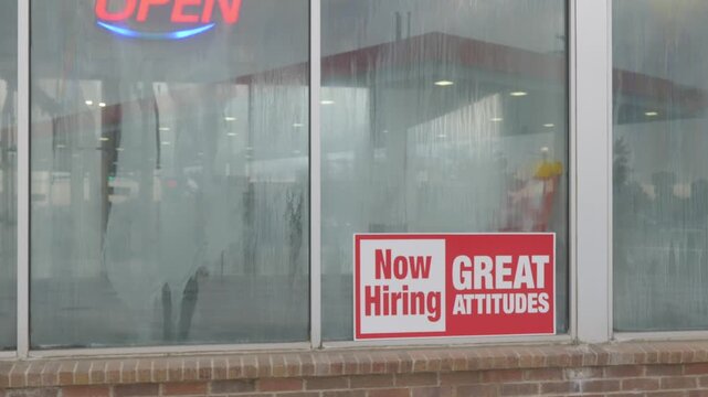 Hiring sign displayed on glass storefront window during daylight hours.