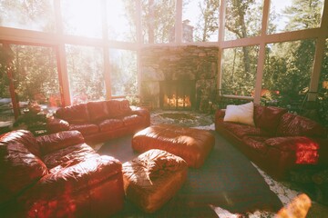 Obraz premium Cherry red leather sofas in a sun-drenched living room with a stone fireplace and forest views for interior design and home decor ideas