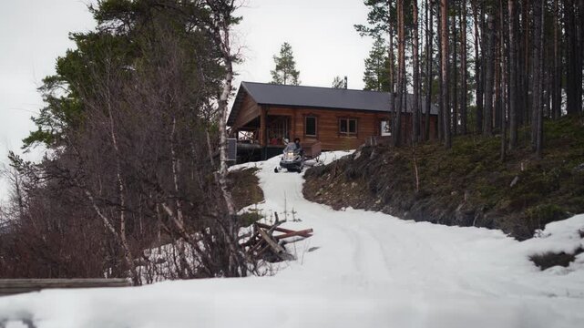 A snowmobile tows a cargo sled away from a remote cabin on a slushy winter track. Ideal for transport, cabin life, travel and outdoor lifestyle content.
