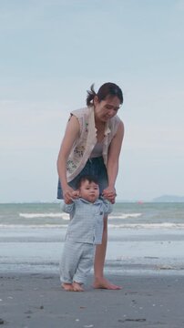 4K Asian mother holding her baby boy's hands, supporting his first steps on the beach.	
