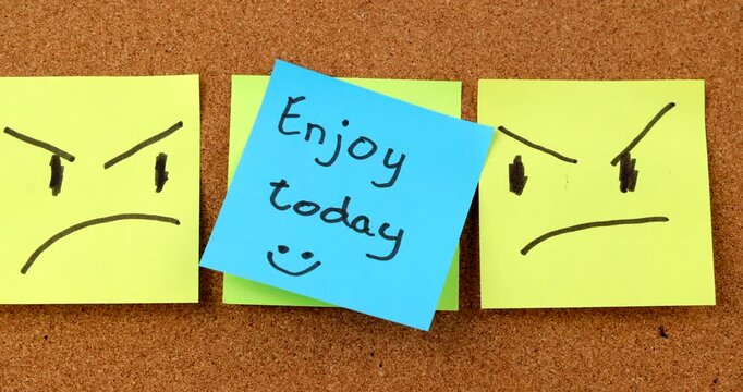 Person sticking blue blank note over a yellow enjoy today smiling memo on brown corkboard, hiding optimism and happy thoughts now.