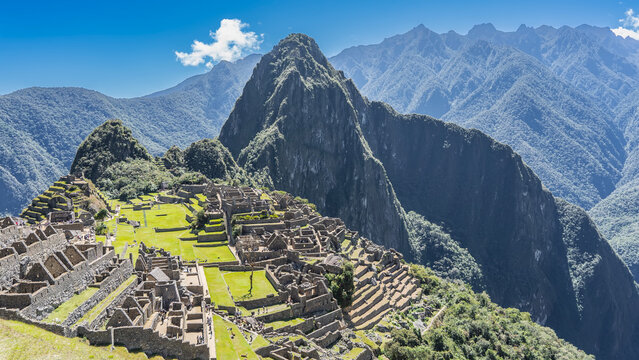 Panorama of the famous Machu Picchu. View from the height. There are dilapidated masonry buildings, green terraces, and stairs on the mountainside.  Huayna Picchu, the Andes mountain range, blue sky.
