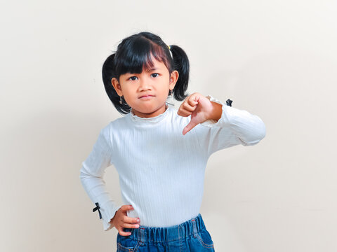 A cute little Asian girl with pigtails hair looking at the camera with a disappointed expression while showing a thumbs down gesture against an isolated background.