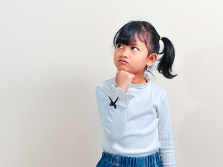 Fototapeta premium A cute little Asian girl with pigtails hair looking up thoughtfully while holding her chin with a hand against an isolated background.