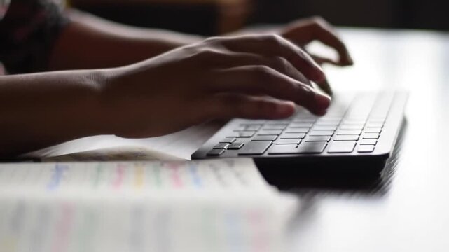 Close-up of hands typing on a computer keyboard with a notebook open