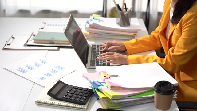 Focused Professional at Work: A dedicated woman in a yellow suit meticulously works on her laptop amidst a desk cluttered with important documents, charts, and office supplies.