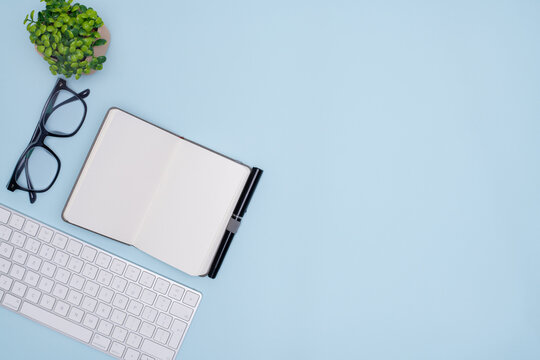 Top View of Office Workspace with Keyboard, Notebook, Pen, Glasses, and Plant on Pastel Blue Background