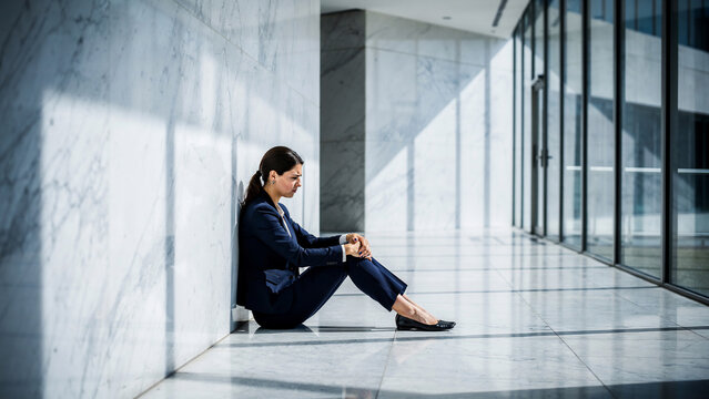 Stressed businesswoman sitting on floor in modern office hallway with marble walls and glass windows, suitable for corporate stress, mental health and business failure themes