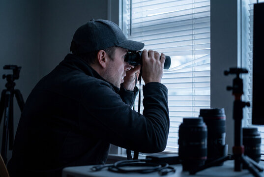 Man with binoculars doing surveillance through window blinds