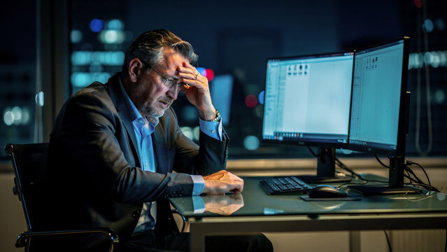 Mature businessman looking stressed at dual monitors in a dark office at night, bokeh city lights, ideal for corporate stress, deadline pressure and overtime work