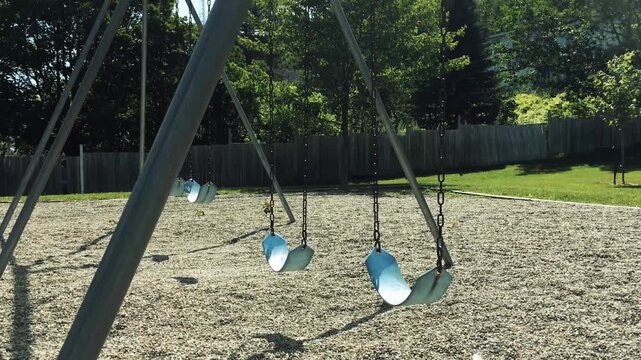 Empty swings moving slowly on playground in summer park at daytime.