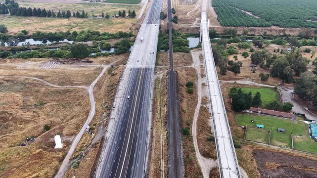 Elevated concrete viaduct under construction for California's High-Speed Rail parallels busy Fresno highway, sparse traffic flows past dry farmland and distant orchards. Aerial drone flyover