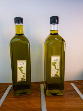 Two bottles of authentic handmade Greek extra virgin olive oil with Corfu island map on the label, displayed on a rustic wooden shelf against a clean white background, souvenir from Greece