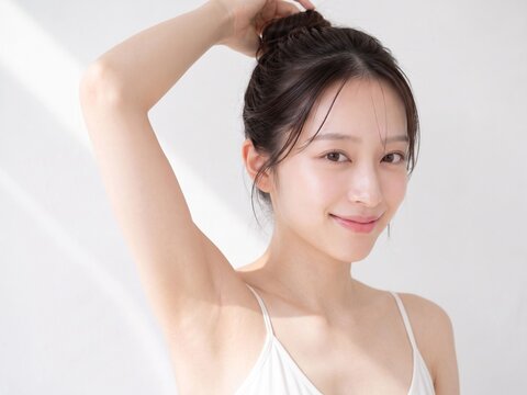 明るい白い背景で片腕を上げてお団子ヘアに手を添え、カメラに向かって穏やかに微笑む若い日本人女性の美容・ボディケア広告向けポートレート
