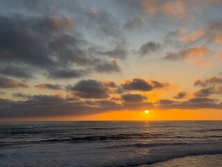 Fototapeta premium Orange and golden hues fill the evening sky and clouds during a dramatic sunset over the Pacific Ocean in Carlsbad, California.