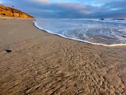 Beautiful landscape of white ocean waves gently rolling onto a wide sandy beach shoreline under a blue sky with soft clouds in Carlsbad, California.
