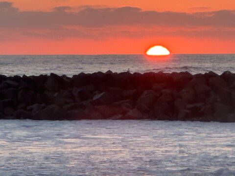 A close up cropped telephoto shot of a vibrant glowing orange sun half way down the horizon behind the silhouette of rock jetty boulders in Carlsbad, California.