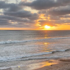 Obraz premium A beautiful vertical composition capturing a radiant golden hour sun setting through dark clouds, casting light on the wet beach sand and waves in Carlsbad.