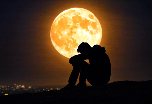Silhouette of a person sitting alone under a glowing supermoon in nighttime landscape