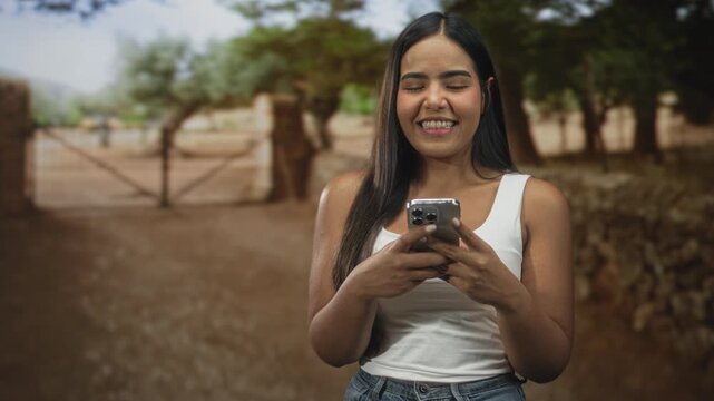 Young woman texting on smartphone with thumbs visible, smiling and holding the phone with both hands on a rural street by a gate and stone wall under trees; happiness.